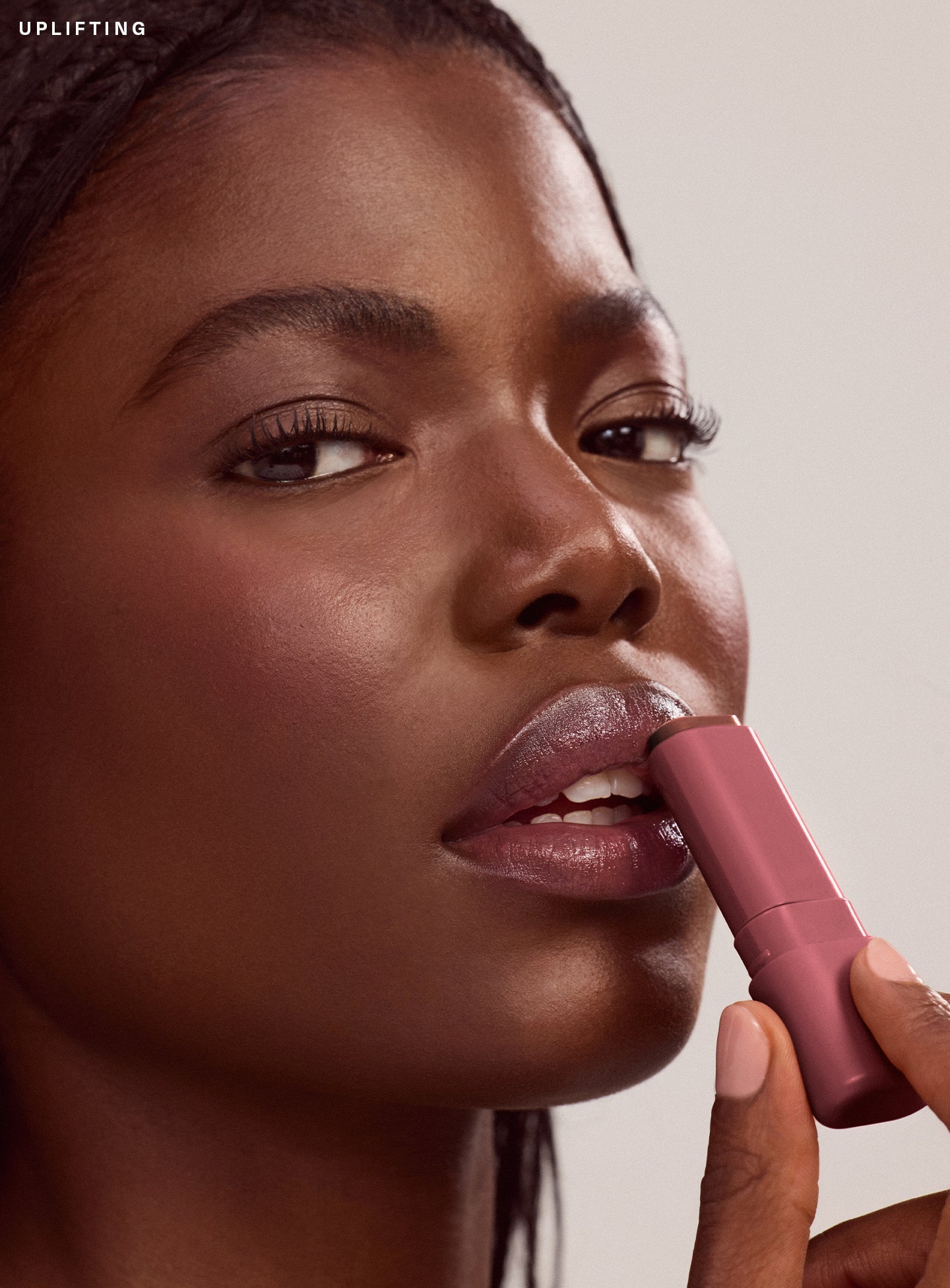 Uplifting| Close-up of model with deep brown skin applying Rare Beauty Find Comfort Lip Butter in shade Uplifting, a deep cool berry color