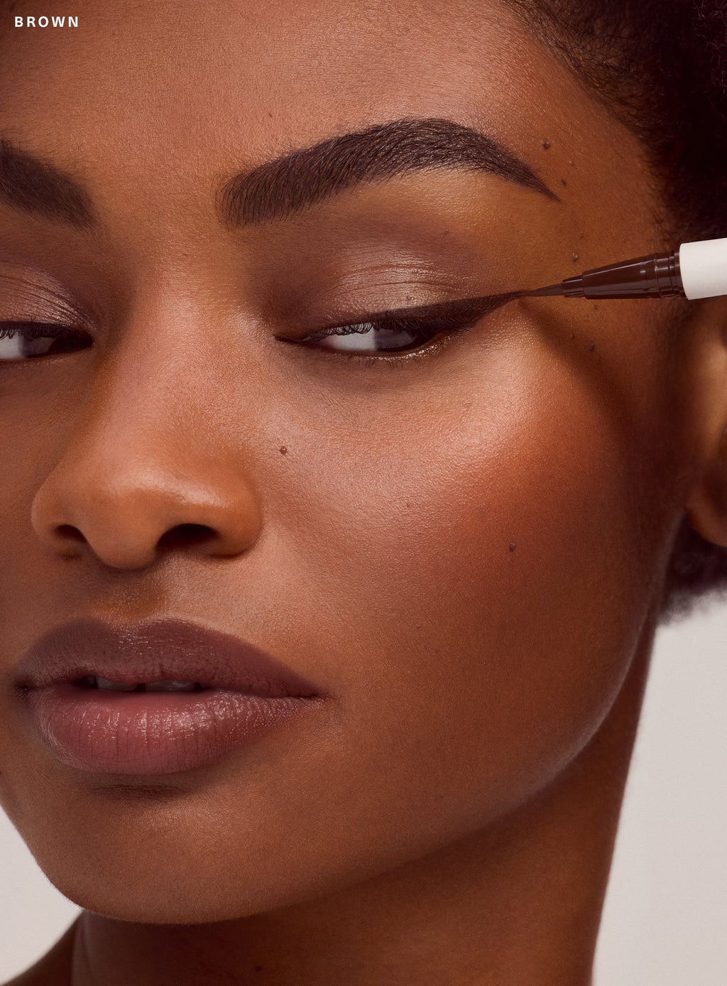Model with deep brown skin tone applying Rare Beauty's Perfect Strokes brown liquid eyeliner