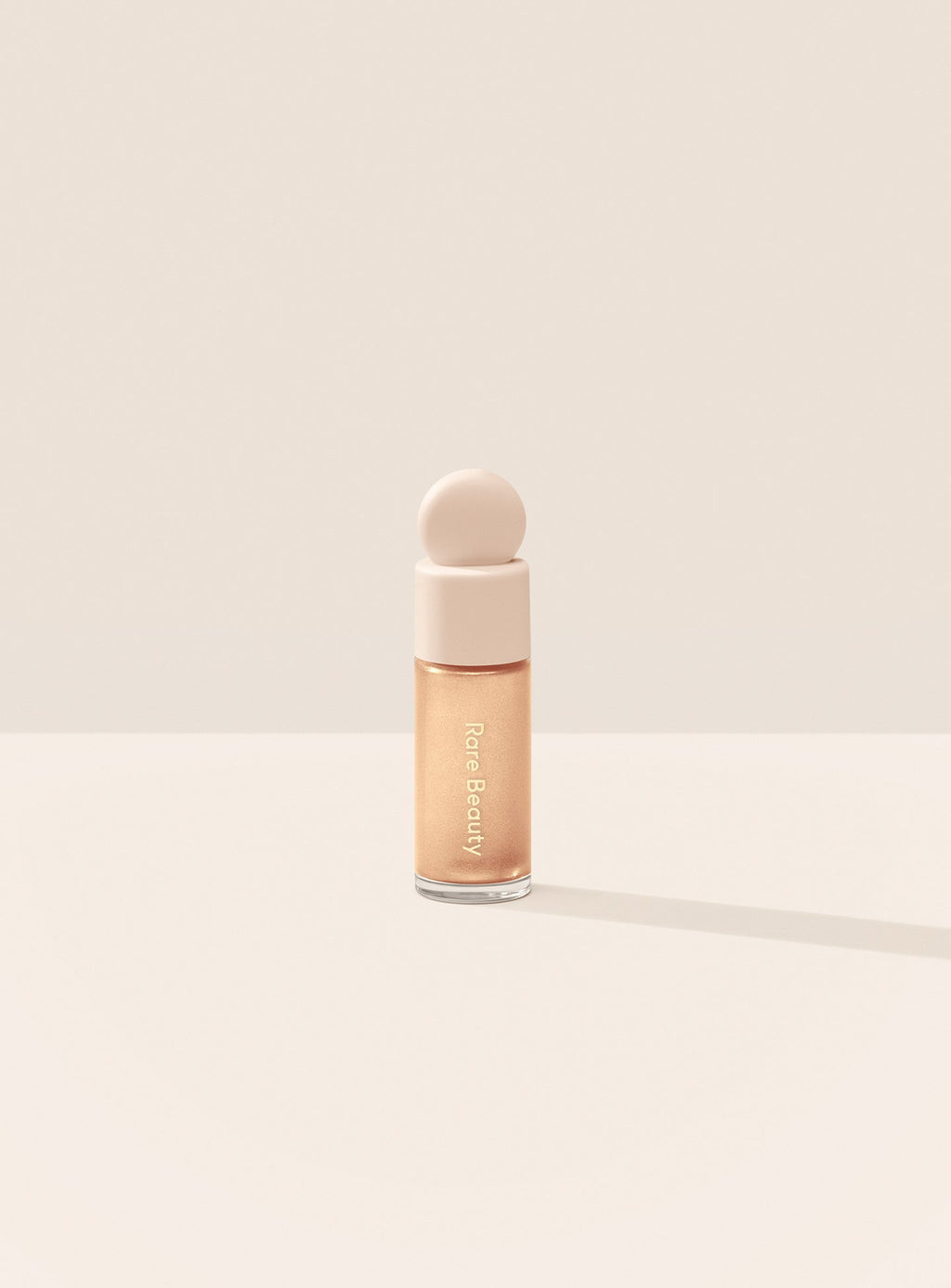 Exhilarate| Rare Beauty's Positive Light Liquid Luminizer Mini in the champagne shade Exhilarate against a neutral background.