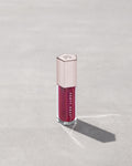 A closed tube of Gloss Bomb Universal Lip Luminizer in the shade Fuschia Flex on a grey background.