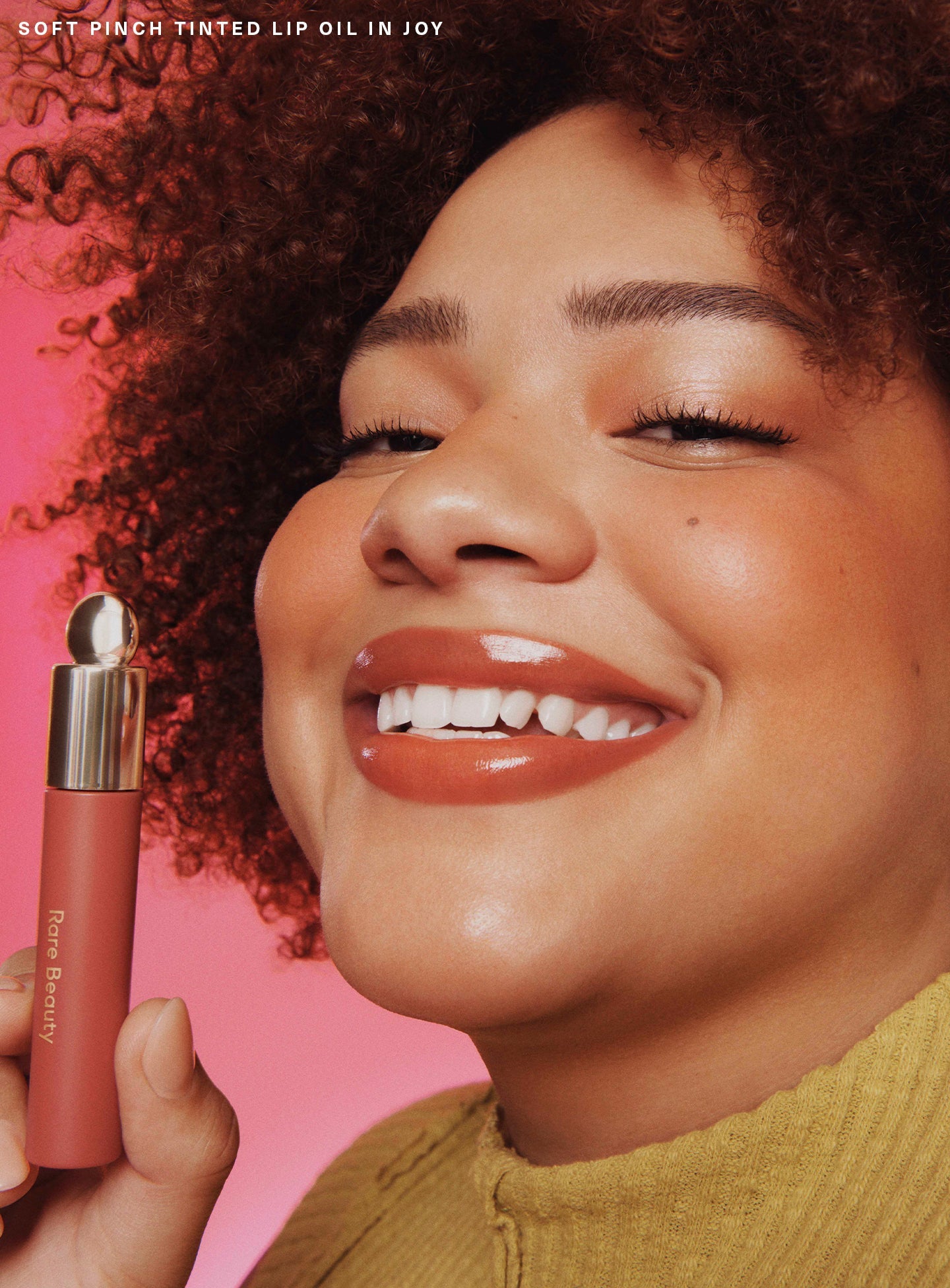 Joy| A model holding Rare Beauty's Soft Pinch Tinted Lip Oil in shade "Joy" and smiling.